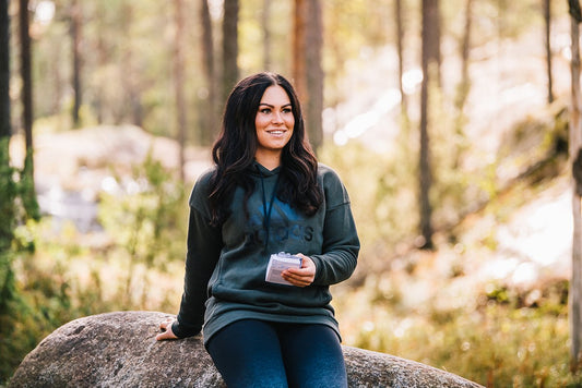 Janette Lepistö sitter på en stein ute i skogen. Hun har langt svart hår, smiler og holder en boks med kosttilskudd med vitamin D.