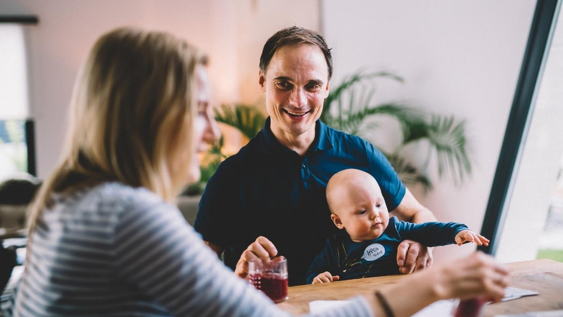 En dame i stripete genser sitter ufokusert i forgrunnen, ved siden av sitter det en smilende mann med en baby i fanget. Kan barnet ditt ha behov for vitaminer?