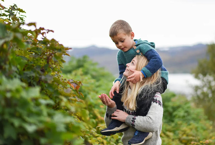 En dame med en liten gutt på skuldrene står mellom flere bærbusker og trær, i bakgrunnen kan en se et vann