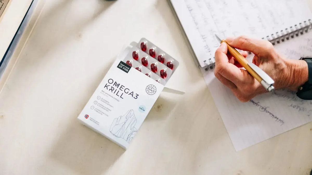 En hvit eske med pillebrett med røde kapsler ligger på en trepult. På boksen står det "Omega 3 Krill" i svart skrift. EN hand holder en penn over en notatblokk på høyre side.