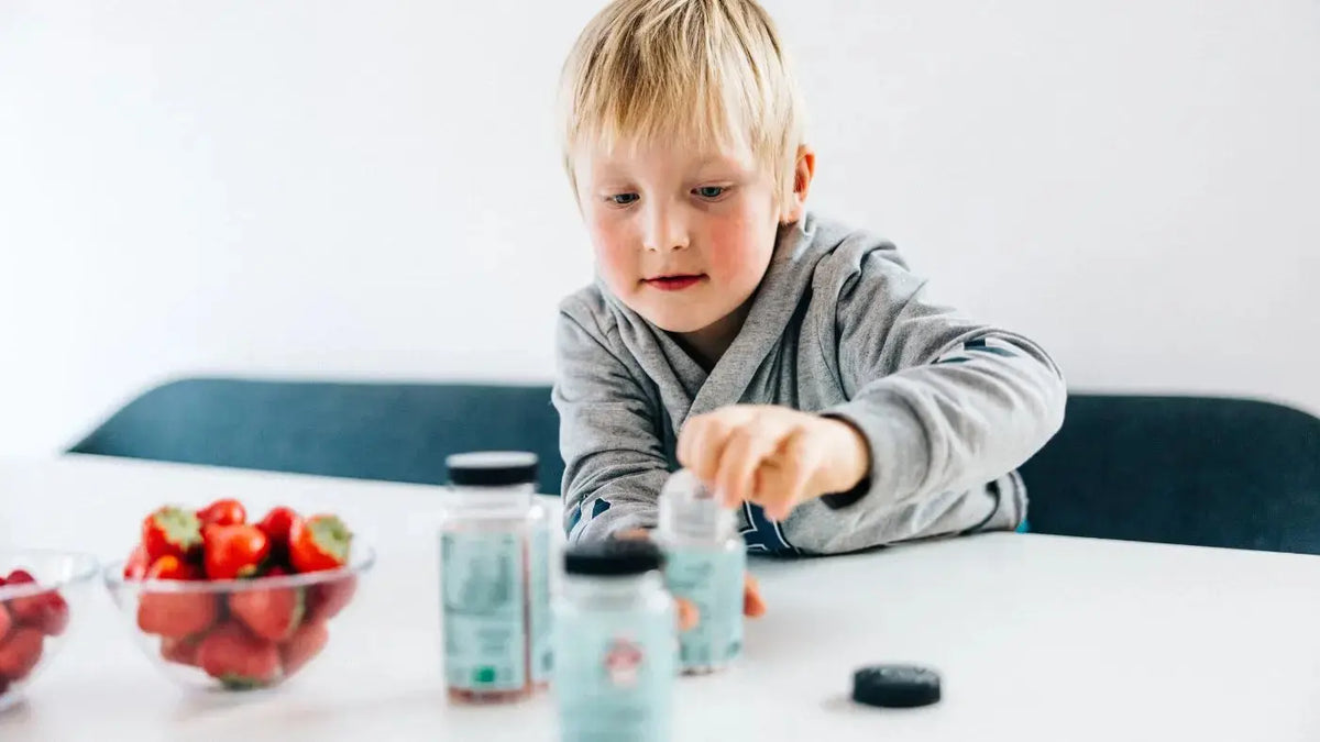 En liten gutt i en grå hettegenser sitter ved et spisebord inne. I hånden har han en grønn boks med kosttilskudd.