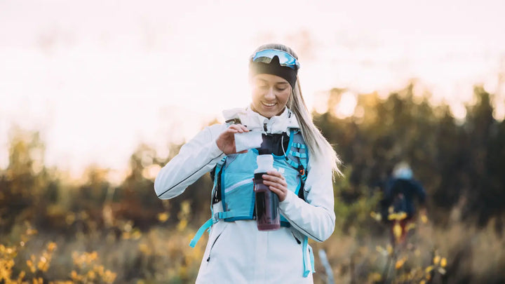 En smilende dame står ute i skogen. Hun har en vindjakke i hvitt og blått på og solbriller på hodet. I hendene står hun med en vannflaske som hun heller kosttilskuddet b12 med tyttebær på.