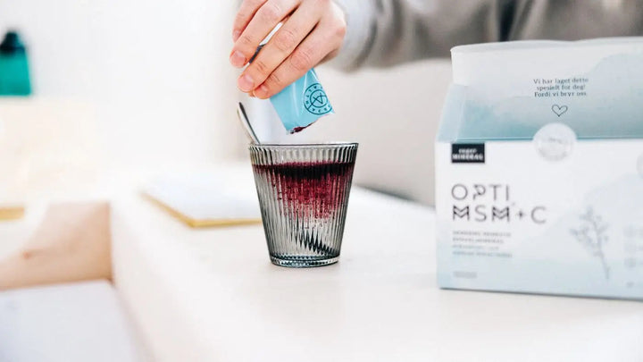 En kvinnehånd med en grå genser heller en blå porsjonspose med OptiMSM+C i et glass med vann og teskje. En pakke med OptiMSM+C (vegan) med svart skrift står på pulten ved siden av glasset. 