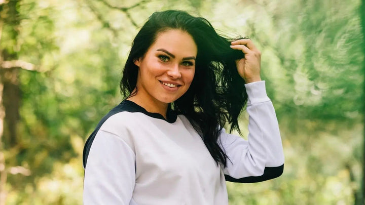 En dame i en hvit genser og med langt, svart hår, ser smilende inn i kameraet. Hun vises fra brystet og opp og står ute i en skog mens hun stryker håret vekk fra ansiktet.