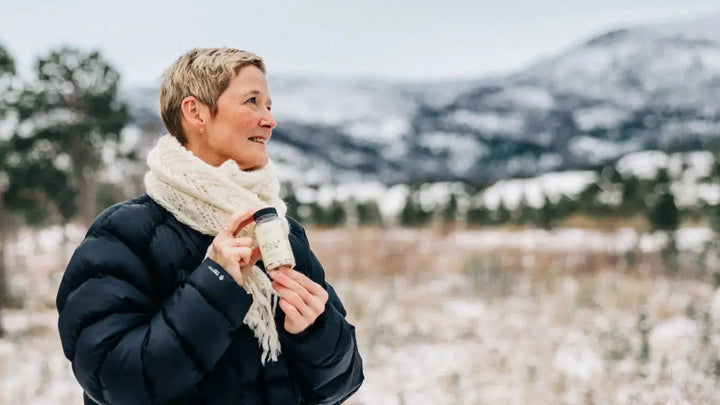 En dame med kort, blondt hår står foran et snødekt område fra hoften og opp. Hun har på en svart boblejakke og et hvitt skjerf, i hånden har hun en boks med kollagen