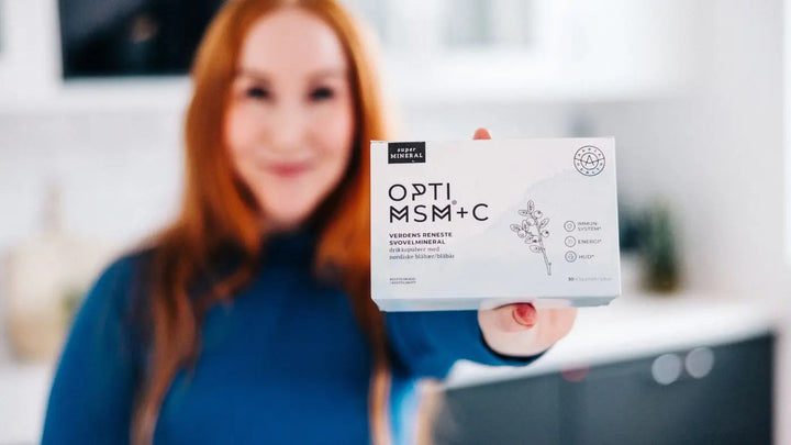 En dame kledt i blått og med langt rødt hår står ufokusert fra brystkassa og opp, mens hun viser fram en boks med OptiMSM + C