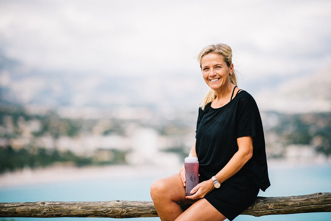 En smilende dame kledt i sort sitter foran en innsjø med en boks med kosttilskudd