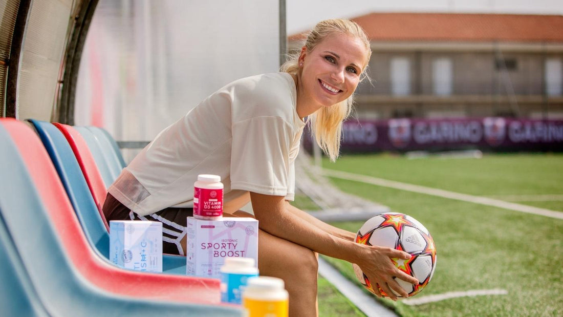På bildet er en smilende Tuija Hyyrynen. Hun sitter på benken ved en fotballbane med en fotball i hendene. I forgrunnen står det flere ulike typer kosttilskudd fra Aarja Health