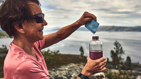 En dame med kort brunt hår, solbriller og en rosa t-skjorte på, heller OptiMSM+C (vegan) pulver i en vannflaske