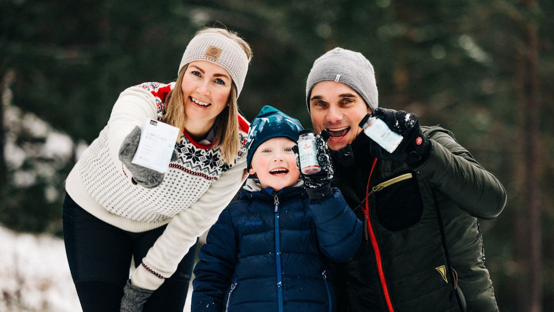 En kvinne, et barn og en mann holder hvert sitt kosttilskudd mens de smiler og ser mot kamera. De er ute i naturen og det er vinter. 