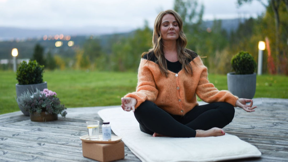 En dame med langt, brunt hår i en oransje strikkejakke, sitter ute på en terrasse i meditasjonsstilling med øynene lukket.