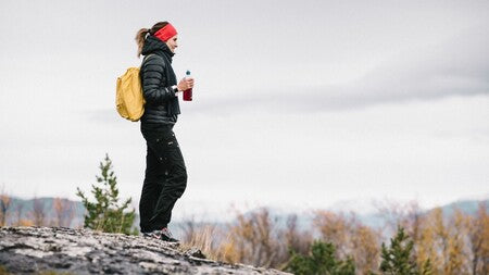 En dame kledt i svart boblejakke, et rødt hårbånd og med en gul sekk på ryggen står oppe på en fjellknaus i profil og skuer ut over området. I den ene hånda har hun en flaske med kosttilskudd i.