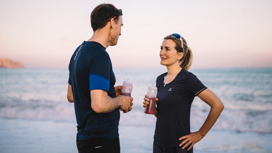 En mann og en dame står og ser på hverandre på en strand. De har på seg svart treningsstøy og holde begge en vannflaske med kosttilskudd.