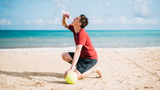 En mann i en rød t-skjorte er på en strand og støtter seg på en ball mens han drikker fra en vannflaske. Artikkelen handler om kosttilskudd på reise.