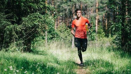 En mann kledt i en rød t-skjorte og svart shorts løper gjennom en skog, i hånden har han en vannflaske