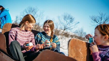 To smilende jenter i strikkegensere sitter utendørs i et vinterlandskap, i hendene holder de bokser med kosttilskudd.
